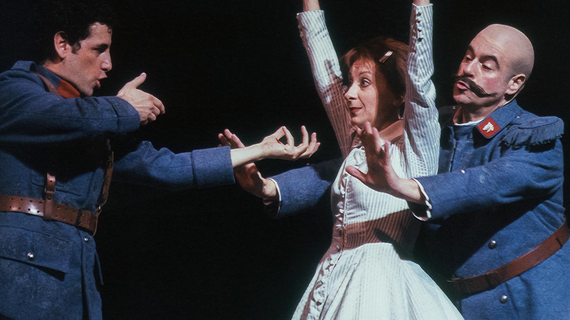 Juan Diego Flórez pointing an imaginary gun at Natalie Dessay, whose hands are raised in the air; a third performer stands behind Dessay with arms pointed out toward Flórez.