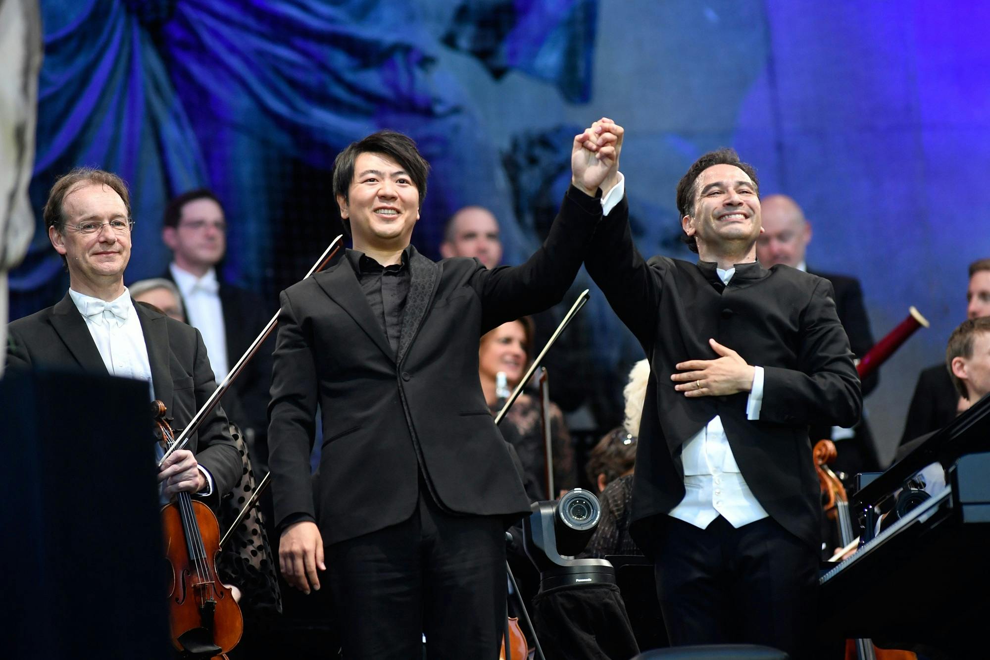 Lang Lang and Andrés Orozco-Estrada holding and raising their hands for applause, with orchestra members standing behind them.