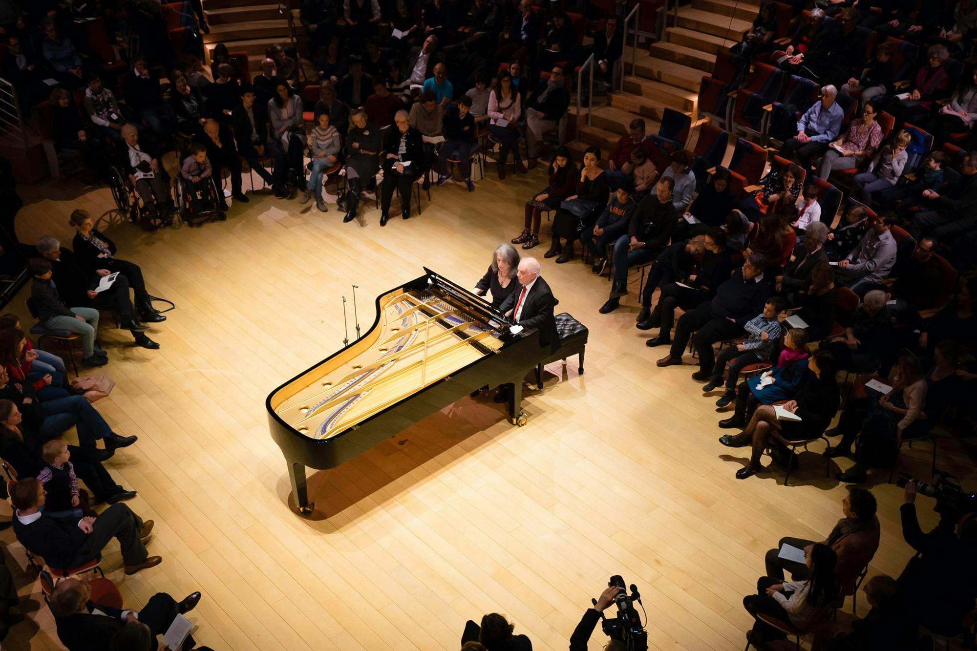 Martha Argerich and Daniel Barenboim playing piano surrounded by an audience.