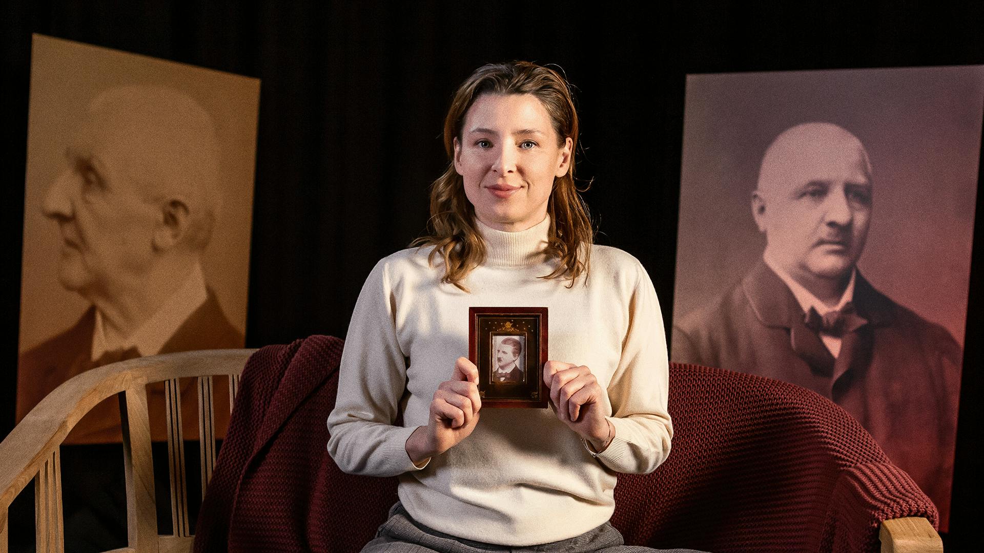 A person holding up a framed photo of Anton Bruckner, and two portraits of Anton Bruckner in the background