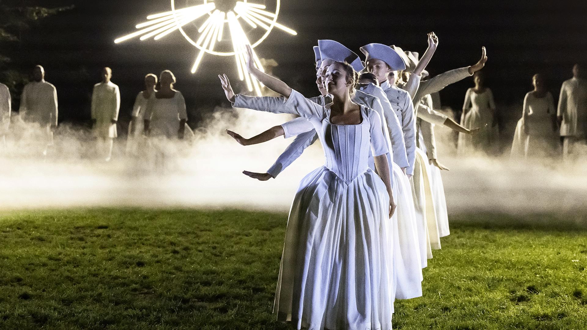 Performers standing in a line on a foggy field, with arms raised