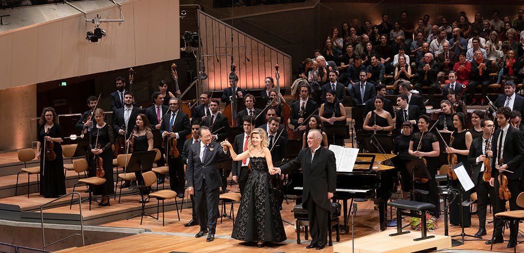 Yo-Yo Ma, Anne-Sophie Mutter, and Daniel Barenboim standing and holding raised hands for applause, with an orchestra standing behind them.