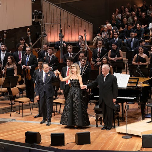 Yo-Yo Ma, Anne-Sophie Mutter, and Daniel Barenboim standing and holding raised hands for applause, with an orchestra standing behind them.