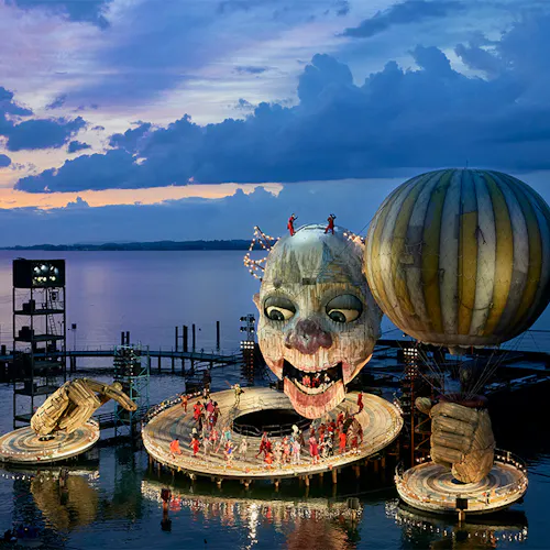 A large structure resembling a clown holding a balloon docked on a waterfront, with a group of people standing on the structure
