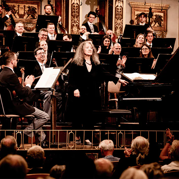 Martha Argerich in front of an orchestra standing for applause.