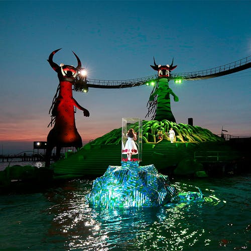 A child standing in a glass box surrounded by a river, looking at bridge held up by three monsters.