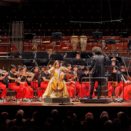 Alisa Weilerstein playing cello on stage, with Rafael Payare conducting NYO2