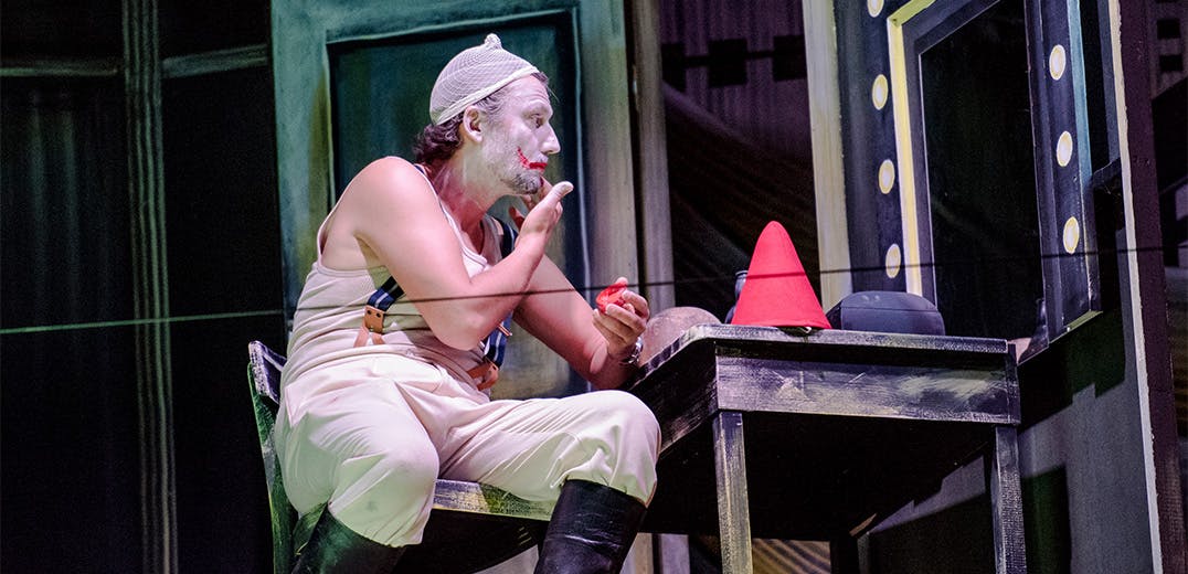 Jonas Kaufmann sitting in a chair on stage in a clown costume applying makeup