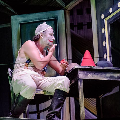 Jonas Kaufmann sitting in a chair on stage in a clown costume applying makeup