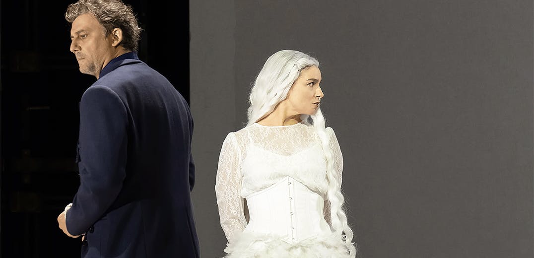 Jonas Kaufmann wearing a suit and Asmik Grigorian wearing a white dress, with their backs facing each other.