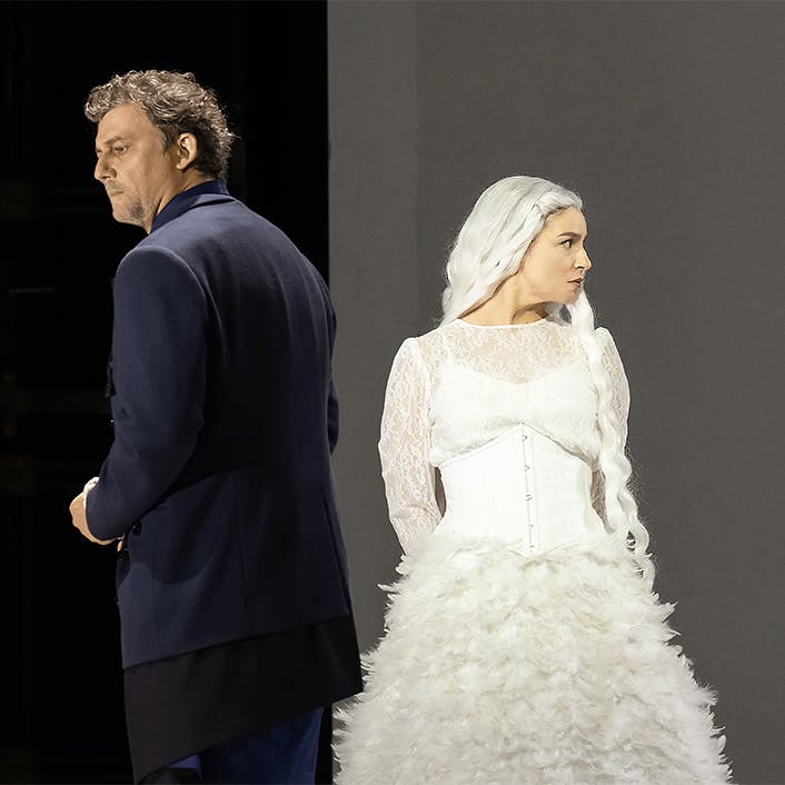 Jonas Kaufmann wearing a suit and Asmik Grigorian wearing a white dress, with their backs facing each other.