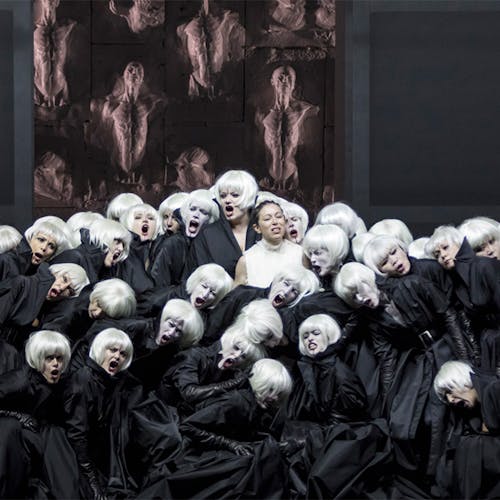 A large group of people with black cloaks and white hair standing in a group, with a person in a white dress and black hair stands in the middle