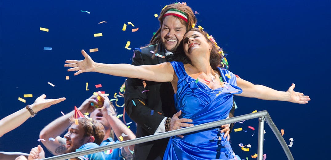 Two people standing together, one with arms wide open, as a crowd throws confetti into the air