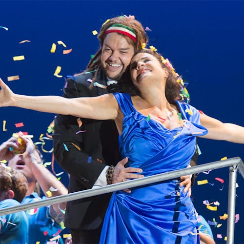 Two people standing together, one with arms wide open, as a crowd throws confetti into the air