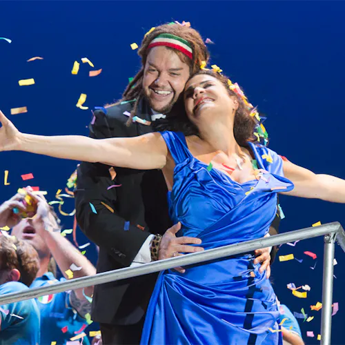 Two people standing together, one with arms wide open, as a crowd throws confetti into the air