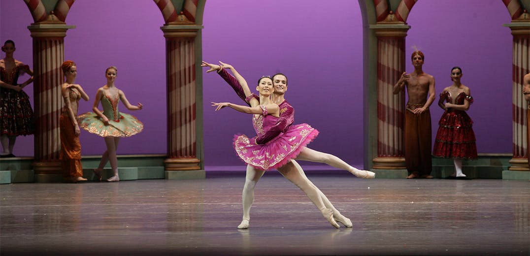 Two dancers on stage, in front of a large column-built exterior, with other people standing nearby