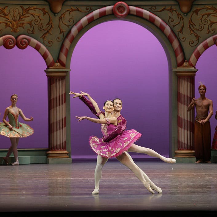 Two dancers on stage, in front of a large column-built exterior, with other people standing nearby
