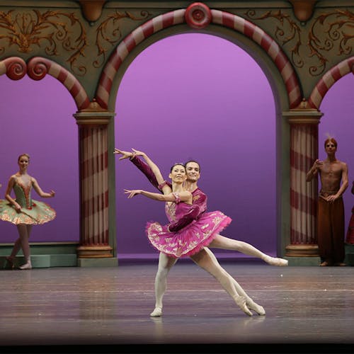 Two dancers on stage, in front of a large column-built exterior, with other people standing nearby