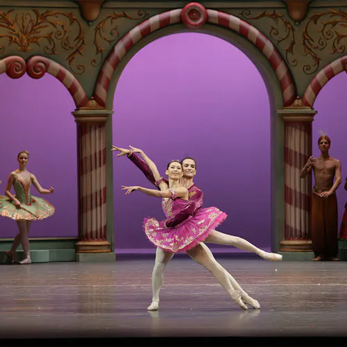Two dancers on stage, in front of a large column-built exterior, with other people standing nearby