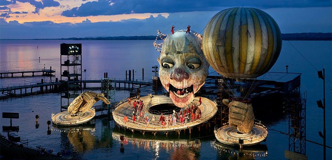 A large structure resembling a clown holding a balloon docked on a waterfront, with a group of people standing on the structure