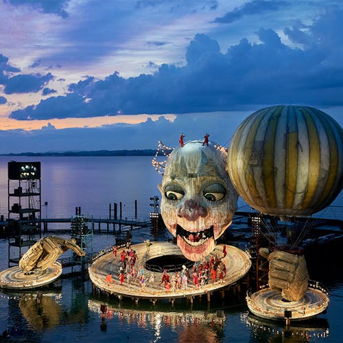 A large structure resembling a clown holding a balloon docked on a waterfront, with a group of people standing on the structure