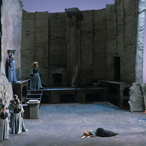 A group of performers standing in front of a large stone structure, with a performer laying down in the middle of the stage