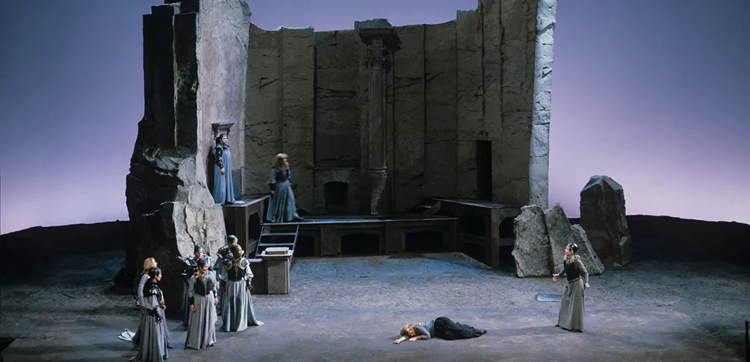 A group of performers standing in front of a large stone structure, with a performer laying down in the middle of the stage