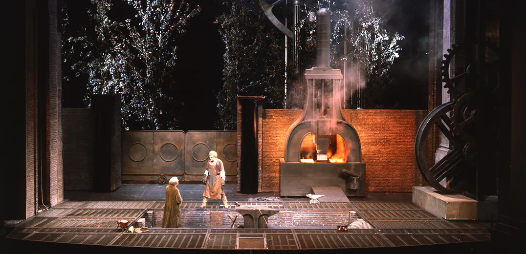 Two performers standing in front of a gate and a large furnace