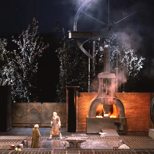 Two performers standing in front of a gate and a large furnace