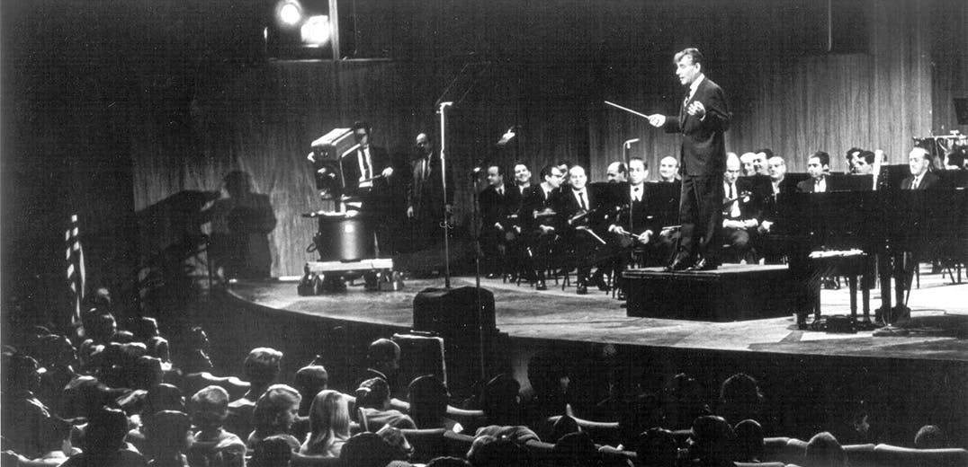 Leonard Bernstein standing on the podium, with an orchestra, facing the audience