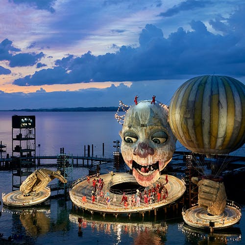 A donut-shaped stage on the water, with a suspended clown head and balloon above.