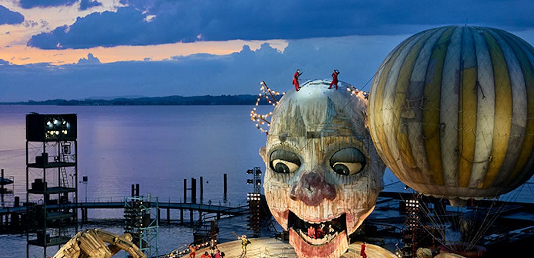 A donut-shaped stage on the water, with a suspended clown head and balloon above.