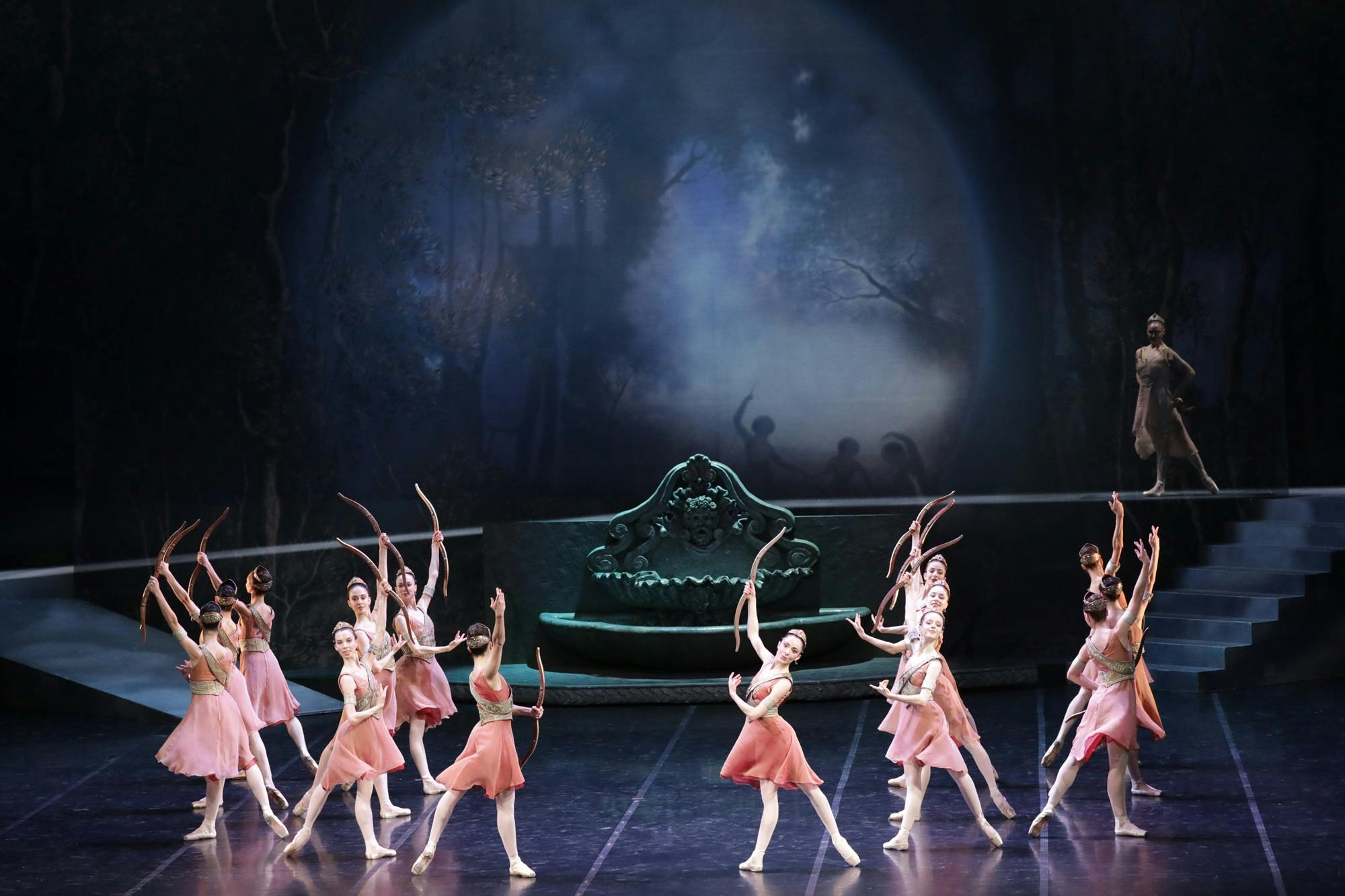 Dancers holding wooden bows, in front of a stone fountain and staircase