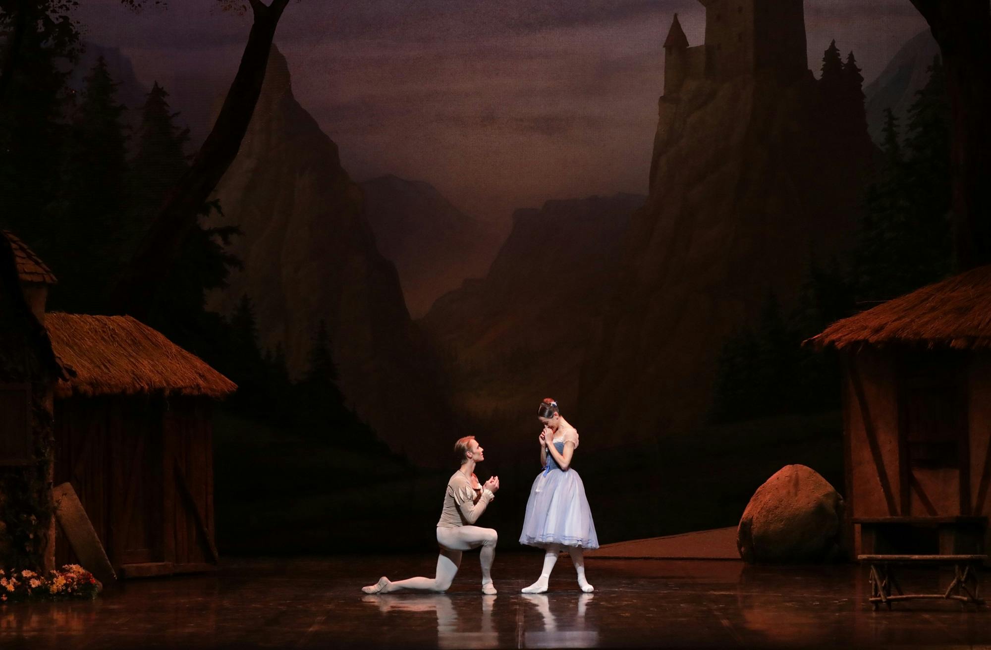 A dancer crouching on one knee in front of another dancer, with straw houses and castles in the background.
