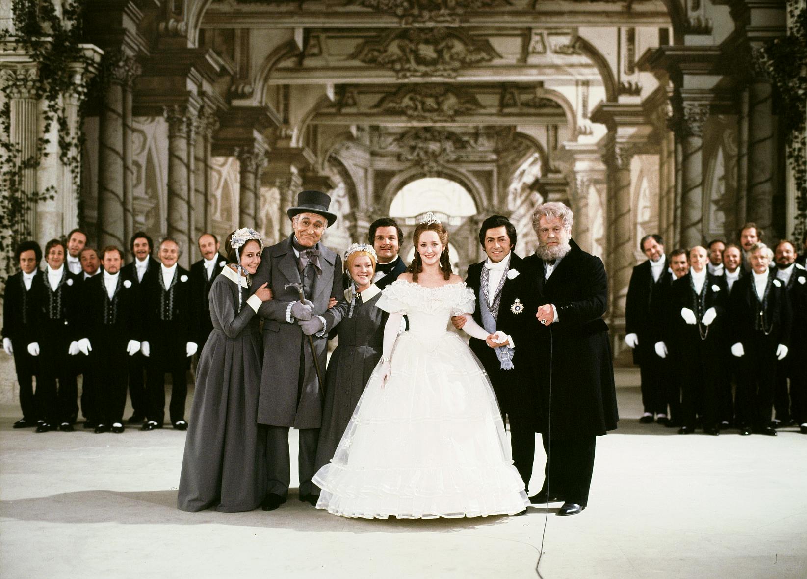 A performer in a white gown surrounded by a group of people in suits and formal clothing.