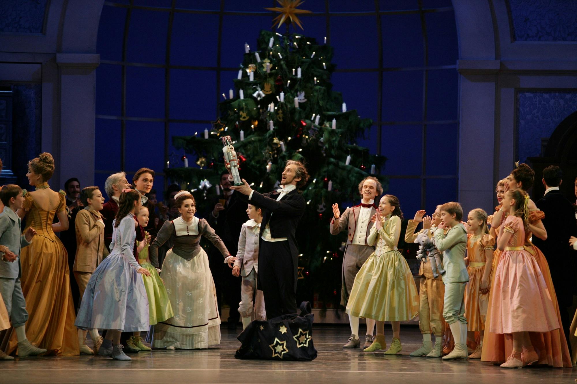 Elegantly dressed ballet performers gathered around a man holding up a large nutcracker figure in front of a decorated Christmas tree.